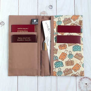 Peut inclure: Un porte-documents de voyage ouvert. Le côté gauche est marron uni, avec des poches contenant un passeport et une petite carte. Le côté droit présente un motif de valises colorées sur fond crème. Un billet d'embarquement et un stylo sont visibles.