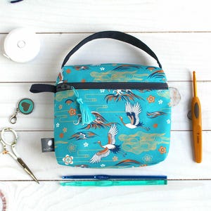 Può includere: Una borsa artigianale turchese con un motivo a gru e nuvole, un manico nero e una cerniera con nappa. Forniture artigianali, tra cui forbici, uncinetti e un metro a nastro, circondano la borsa.