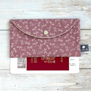 May include: A mauve fabric passport holder with a dragonfly pattern. The holder has a flap closure with a button and a beige trim. A passport is partially visible inside. The holder has a small tag that says "cotton cat".