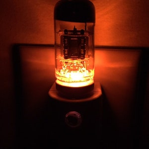 May include: A vintage-style night light with a glowing orange vacuum tube. The light is plugged into a standard electrical outlet.