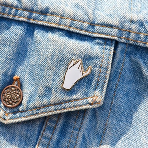 May include: A white enamel pin shaped like a hand making a pinky promise gesture. The pin is attached to a blue denim jacket.
