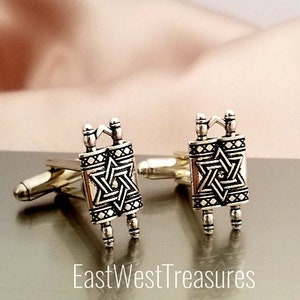 May include: A pair of silver cufflinks featuring a detailed design of a Torah scroll with a Star of David. The cufflinks are on a silver background.