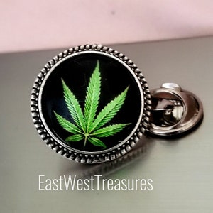 May include: A silver pin with a black background and a green marijuana leaf design. The pin has a decorative border.