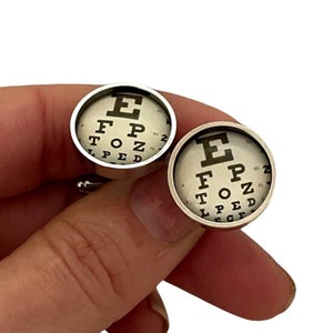 May include: A pair of silver cufflinks with a white background and black eye chart design. The eye chart features the letters "E", "F", "P", "T", "O", "Z", and "P" in descending size.