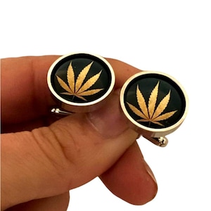 May include: A pair of silver cufflinks with a black background and a gold marijuana leaf design.