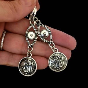 May include: Silver earrings with a dangling charm featuring the Arabic word for God, "Allah", surrounded by an intricate design. The top of the earring features a silver eye charm.