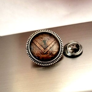 May include: A silver-toned round pin with a brown shield design. The shield has a textured surface and a chevron pattern. The pin has a beaded edge.