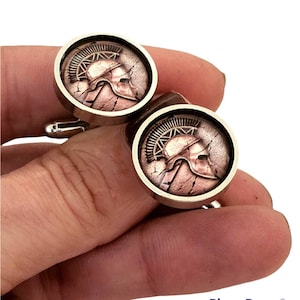 May include: A pair of silver cufflinks with a round, copper-toned design featuring a detailed image of a Spartan helmet.