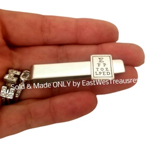 May include: Silver tie clip with a small eye chart charm. The eye chart reads "E, F, P, T, O, Z, L, P, E, D".
