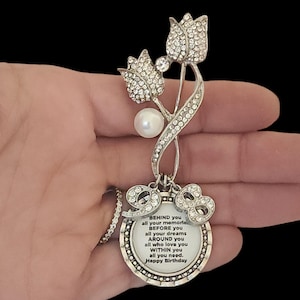 May include: Silver tone brooch with two crystal-encrusted tulips and a pearl. The brooch has a round charm with a message that reads "BEHIND you all your memories BEFORE you all your dreams AROUND you all who love you WITHIN you all you need. Happy Birthday"
