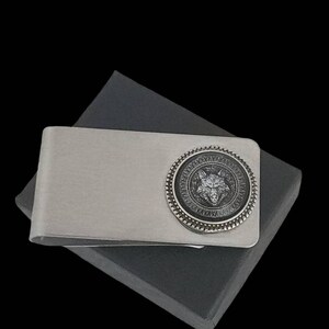 May include: Silver money clip with a circular design featuring a wolf head in the center. The wolf head is surrounded by a decorative pattern. The money clip is on a gray background.