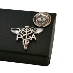 May include: Silver-toned pin with a caduceus symbol and the letters "PSA" on a black background. The pin is in a black box with a silver-toned pin back.