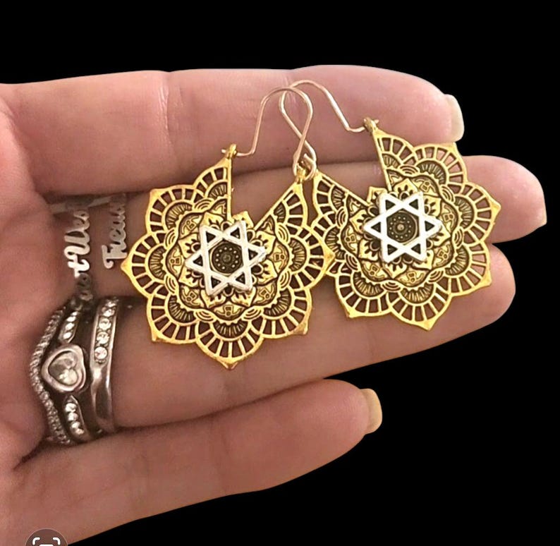 May include: Gold-toned filigree earrings with a floral design and a silver Star of David in the center. The earrings have a hook closure. The earrings are held in a hand.