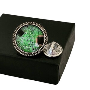 May include: A silver pin with a round green and black circuit board design. The pin is in a black box.