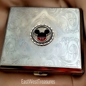May include: Silver cigarette case with an engraved floral design and a Mickey Mouse emblem in the center.