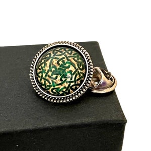 May include: A silver lapel pin with a green and white Celtic knot design. The pin has a decorative border and a safety clasp.