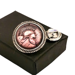 May include: A silver-toned lapel pin with a round, glass-like design featuring a raised image of a Spartan helmet in a bronze color. The helmet is set against a pink and white marble-like background. The pin is in a black box.