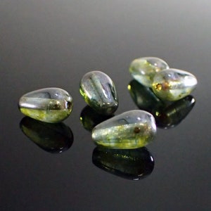 May include: A group of six iridescent green glass beads in the shape of teardrops. The beads have a pearlescent finish and are scattered on a black surface.