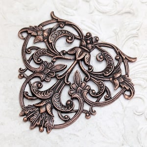 May include: Ornate, copper-colored metal embellishment with a floral and scroll design. The decorative piece features detailed leaves, flowers, and swirling patterns. The symmetrical design is ideal for crafting and decorative projects.