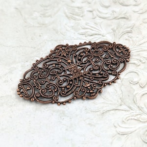 May include: A copper filigree design with intricate swirls and floral patterns. The design is oval shaped and has a delicate, ornate look.