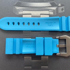 Puede incluir: Correa de reloj azul brillante con hebilla de acero inoxidable. La correa est&aacute; hecha de un material flexible y presenta el texto "Made in Italy" y "OFFICIAL PANERAI". La correa tiene m&uacute;ltiples orificios para ajuste.