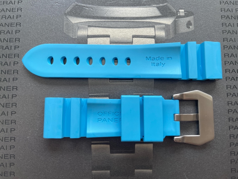 Puede incluir: Correa de reloj azul brillante con hebilla de acero inoxidable. La correa est&aacute; hecha de un material flexible y presenta el texto "Made in Italy" y "OFFICIAL PANERAI". La correa tiene m&uacute;ltiples orificios para ajuste.