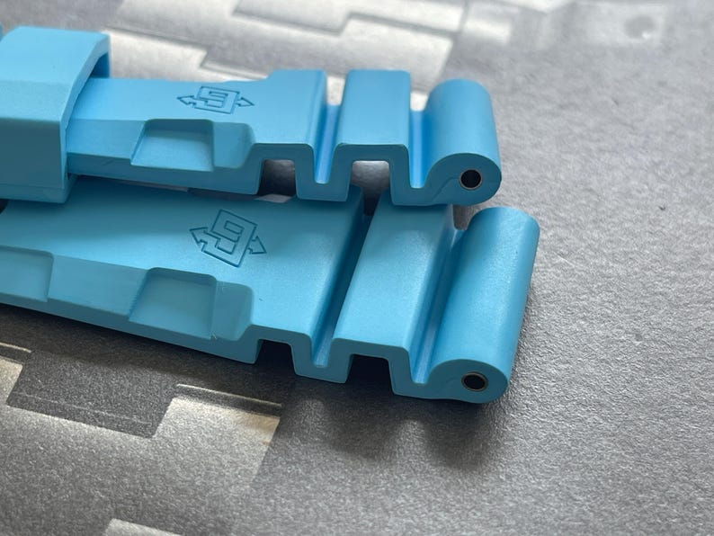 Puede incluir: Primer plano de una correa de reloj azul claro. La correa est&aacute; hecha de un material flexible y presenta una serie de recortes rectangulares y un cierre. La correa tiene el n&uacute;mero "9" y un s&iacute;mbolo de flecha impreso.