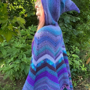 May include: A hooded crochet cape featuring a chevron pattern in various shades of blue, purple, and violet. The garment includes a pointed hood and a textured knit construction with horizontal stripes of varying widths and color gradients throughout.