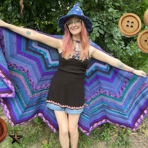May include: A large, star-shaped crochet shawl featuring concentric chevron patterns in shades of blue, purple, and magenta. The shawl is paired with a matching blue crochet pointed hat and three round, four-hole wooden buttons.