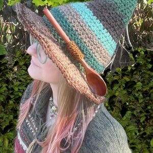 May include: A handmade crochet witch hat with a wooden spoon attached. The hat features stripes of teal, grey, and green. The brim is a darker brown. The person is wearing a green sweater with a patterned design.