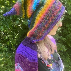 May include: A crocheted hood and shoulder wrap featuring a vibrant rainbow color palette with stripes of red, orange, yellow, green, blue, and purple. The hood has a pointed, elongated shape, and the attached wrap covers the shoulders in textured yarn.
