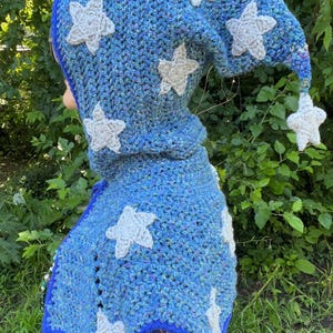 May include: A blue crochet hooded cowl featuring a pointed top and multiple white star-shaped appliqu&eacute;s. The yarn has a variegated texture with flecks of different colors, and the edges are finished with a solid blue border trim.