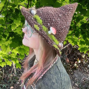 May include: A brown, pointed crochet hat decorated with green moss, small mushrooms, and acorns. The hat has a speckled texture and a whimsical, fairy-tale aesthetic. The hat is worn outdoors, with green foliage in the background.