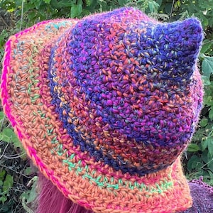 May include: A colorful crocheted witch hat with a pointed top and a wide brim. The hat is made with a variety of shades of purple, pink, orange, and green yarn.