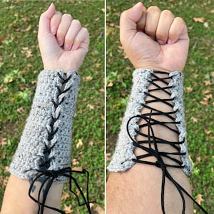 Könnte beinhalten: Graue Häkel-Handgelenkmanschetten mit schwarzer Schnürung. Die Manschetten werden am Handgelenk einer Person gezeigt, wobei die schwarze Schnürung ein dekoratives Muster bildet. Die Manschetten sind aus strukturiertem Garn gefertigt und haben eine neutrale Farbe.