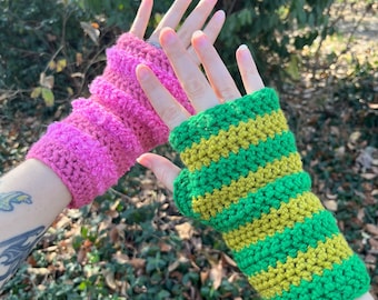 May include: Two handmade crochet fingerless gloves. One is bright pink, the other is green with yellow stripes. The gloves are displayed against a backdrop of green foliage. The crochet stitch is visible, adding texture to the design.