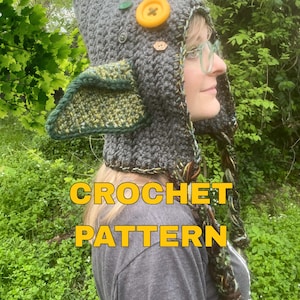 Puede incluir: Un gorro de ganchillo gris con la parte superior puntiaguda, orejeras y lazos trenzados. El gorro está decorado con un botón amarillo grande y botones verdes y marrones más pequeños. El texto "CROCHET PATTERN" se muestra en amarillo.