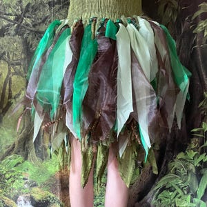 May include: A green and brown fairy skirt with a matching green top. The skirt is made of layered fabric in shades of green, brown, and white. The top is a textured, olive green color. The skirt has a whimsical, fantasy theme.