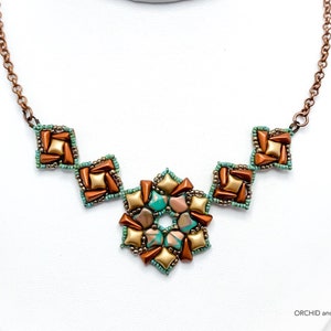 May include: A handmade necklace with a copper-toned chain and pendant. The pendant has a central floral design with turquoise, gold, and brown accents, surrounded by square elements in similar colours. The necklace is displayed on a white surface.