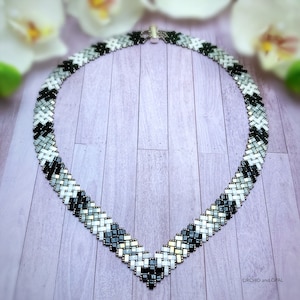 May include: A black, white and silver beaded necklace with a chevron pattern. The necklace is on a light purple wooden surface.