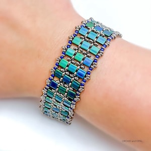 Puede incluir: Un brazalete de cuentas con un patrón geométrico en tonos azules y verdes. El brazalete está hecho con cuentas pequeñas cuadradas y cuentas plateadas.