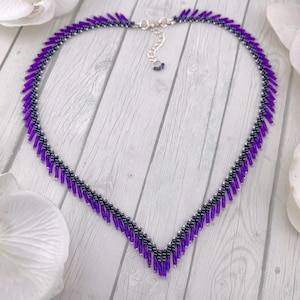 May include: A beaded necklace featuring a V-shaped design composed of vibrant purple bugle beads and small metallic black seed beads. The necklace includes a silver-toned metal chain extender with a lobster claw clasp at the top center.