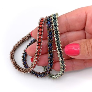 May include: Three beaded bracelets featuring intricate woven patterns. One bracelet is bright orange, one is deep purple, and one is dark green. Each bracelet is constructed from small, faceted, metallic-finish beads arranged in a dense, textured, cylindrical design.