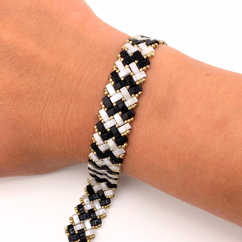 K&ouml;nnte beinhalten: Ein gewebtes Armband mit einem sich wiederholenden diagonalen Muster aus rechteckigen schwarzen und wei&szlig;en Perlen. Die Kanten des Armbands sind mit kleinen, runden metallisch goldenen Perlen besetzt, die das geometrische Design durchgehend betonen.