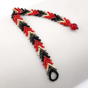 Könnte beinhalten: Ein Perlenarmband mit einem Chevron-Muster aus kleinen roten, schwarzen und cremefarbenen Saatperlen. Das Armband verfügt über eine einzelne runde rote Perle an einem Ende und eine kreisförmige schwarze Perlenschlaufe am anderen Ende.