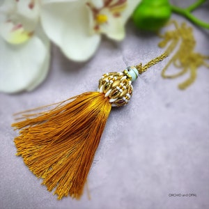 May include: A gold chain necklace with a tassel pendant. The tassel is made of a golden brown fabric and the pendant is a round bead with white and gold accents.