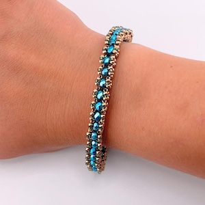 Op de afbeelding: Een kralenarmband met een centrale rij gefacetteerde, metallic turquoise blauwe kralen, geflankeerd door twee rijen kleinere, ronde, metallic gouden kralen. De kralen zijn in een gestructureerd, herhalend patroon geweven tot een band.