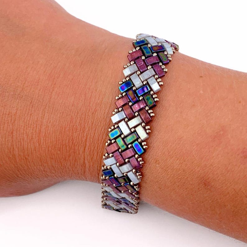 K&ouml;nnte beinhalten: Ein gewebtes Armband mit einem sich wiederholenden Muster aus rechteckigen Perlen in schillerndem Blau, tiefem Violett und perlmuttartigem Wei&szlig;. Die Perlen sind in einem diagonalen Fischgr&auml;tenmuster angeordnet und durch kleine goldfarbene Perlen verbunden.