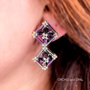 May include: A pair of dangle earrings with a geometric design. The earrings are made of silver-toned metal beads and feature a purple iridescent crystal in the center of each square.
