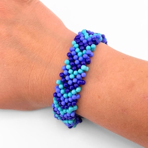 Puede incluir: Una pulsera de cuentas con un patrón tejido denso de pequeñas cuentas redondas en varios tonos de azul, incluyendo azul marino oscuro, bígaro medio y turquesa brillante, dispuestas en un diseño cilíndrico texturizado.
